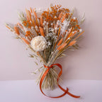 A dried floral arrangement featuring orange wheat, orange babies breath, a large white peony-like sola flower, and a rust-orange ribbon tied at the base, photographed against a pale pink wall and white surface.