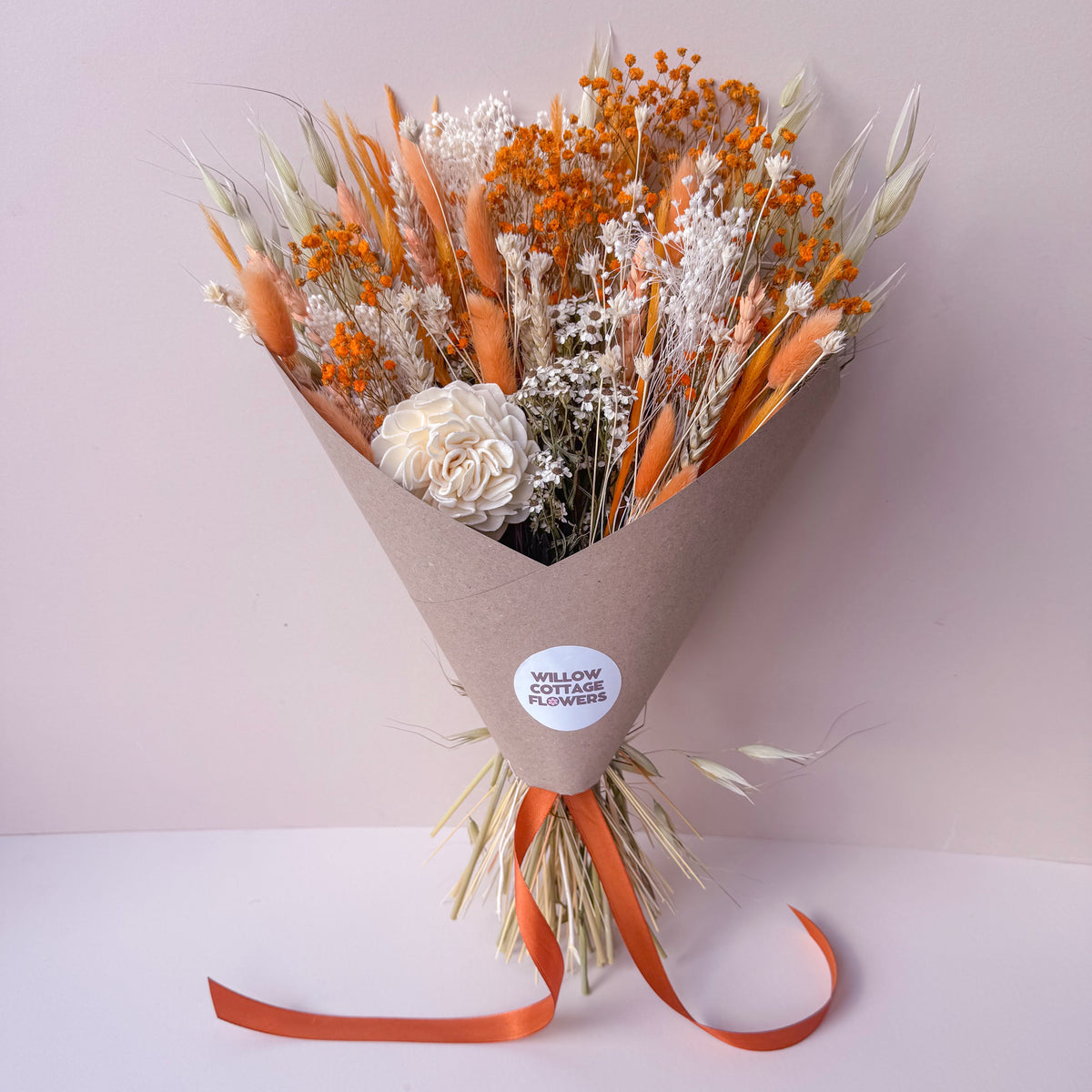 A dried flower bouquet wrapped in brown kraft paper with an orange ribbon and a round white sticker that reads 'WILLOW COTTAGE FLOWERS'. The bouquet features orange, beige, and white dried elements, set against a soft pink background.