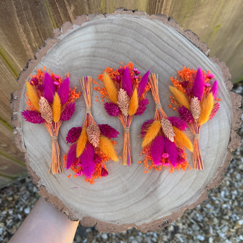 Five dried flower buttonholes with bright magenta, orange and coral botanicals arranged on a round wood slice, held by a hand against a wooden fence and gravel background.