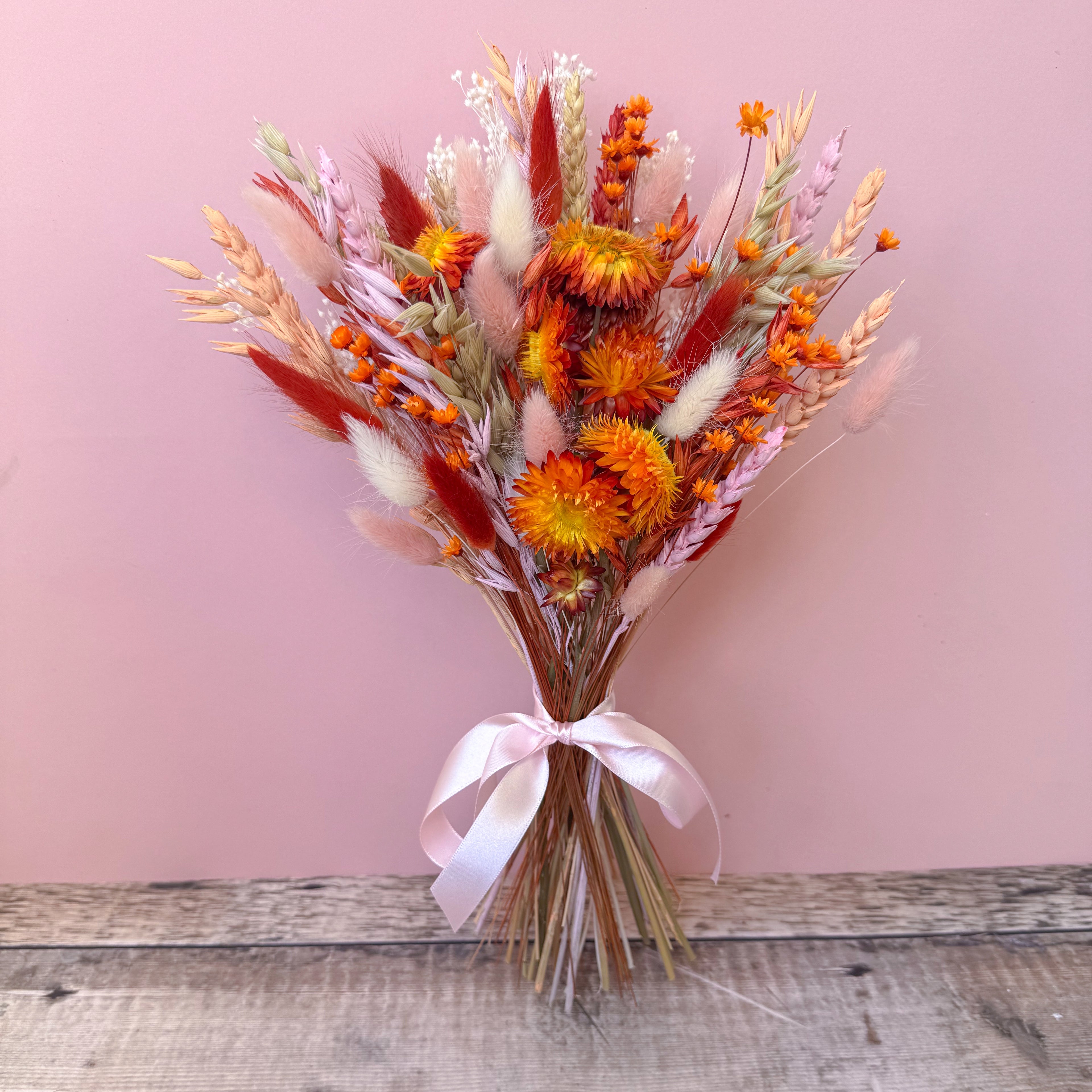 A dried flower bouquet with orange, red, pink, and beige botanicals tied with a pale pink ribbon, set against a dusty pink wall and rustic wooden surface.