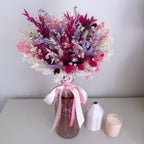 Bouquet of dried flowers in a pink glass vase with pink ribbons on a white surface.