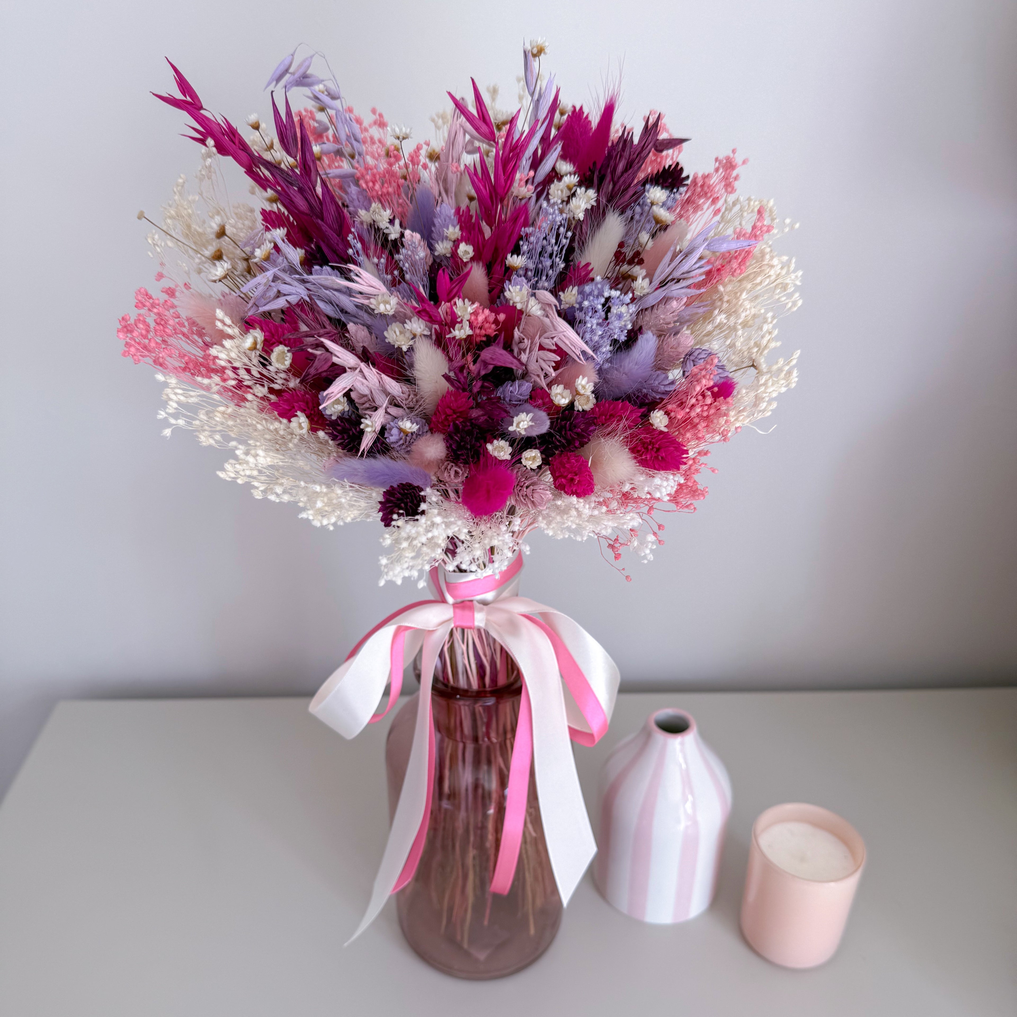 Bouquet of dried flowers in a pink glass vase with pink ribbons on a white surface.