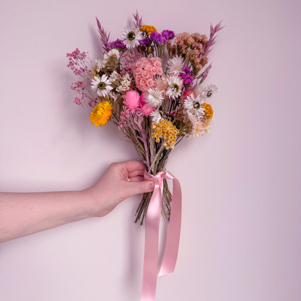 Bouquet of colorful flowers held by a hand with a pink ribbon against a light pink background