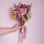 Bouquet of colorful flowers held by a hand with a pink ribbon against a light pink background