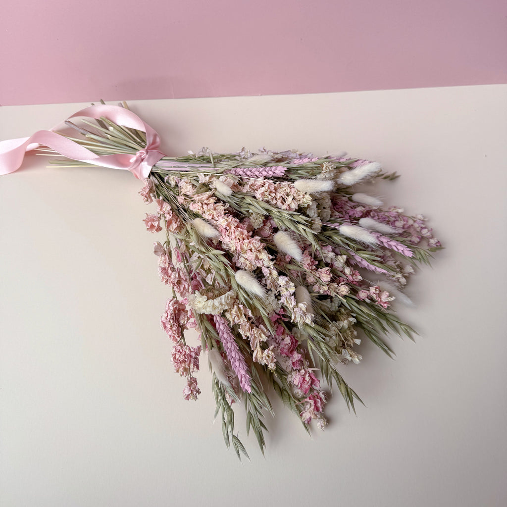 A dried flower bouquet with pink and white blossoms, green stems, and a light pink ribbon, arranged on a two-tone pink and cream background.