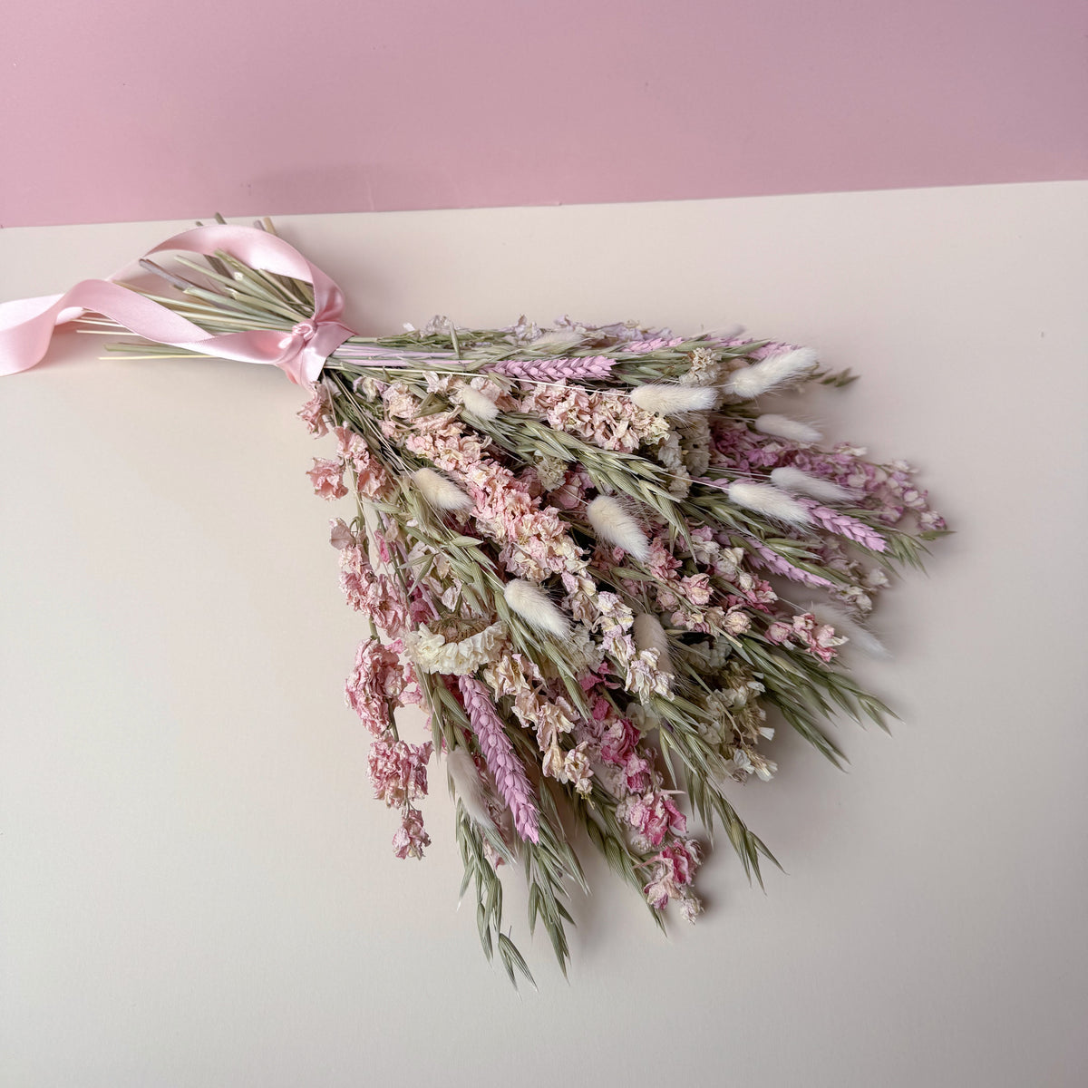 A dried flower bouquet with pink and white blossoms, green stems, and a light pink ribbon, arranged on a two-tone pink and cream background.