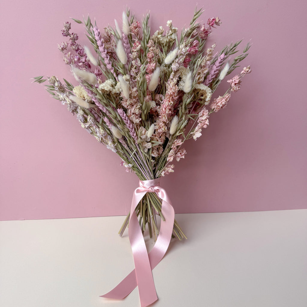 A dried flower bouquet with pink, white, and beige botanicals tied with a pink satin ribbon, set against a split pink and white background.