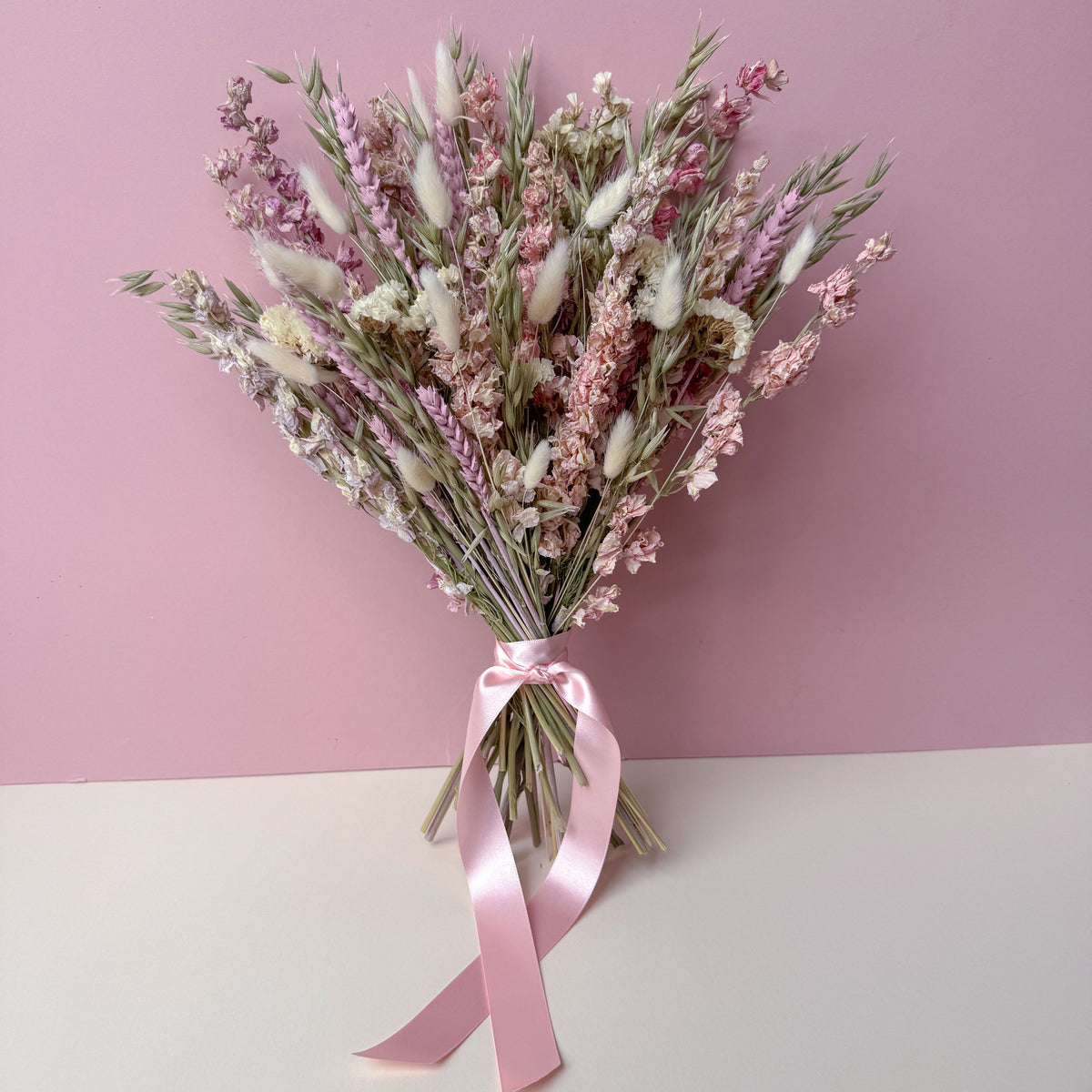 A dried flower bouquet with pink, white, and beige botanicals tied with a pink satin ribbon, set against a split pink and white background.