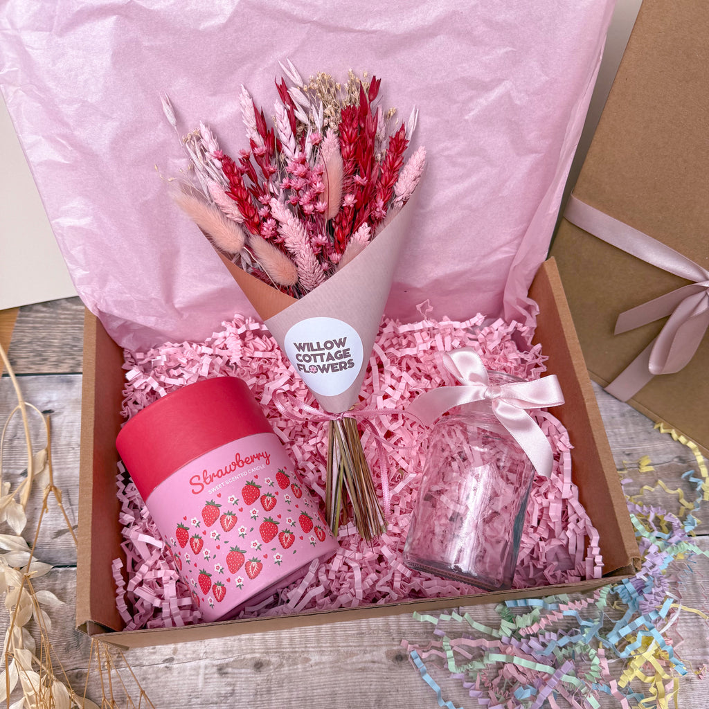 A gift box filled with pink shredded paper contains a bouquet of dried pink and red flowers, a pink strawberry-themed candle, and a small clear glass jar tied with a pale pink ribbon. The box is lined with pink tissue paper and sits on a rustic wooden surface.