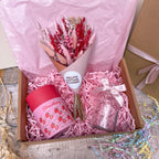 A gift box filled with pink shredded paper contains a bouquet of dried pink and red flowers, a pink strawberry-themed candle, and a small clear glass jar tied with a pale pink ribbon. The box is lined with pink tissue paper and sits on a rustic wooden surface.