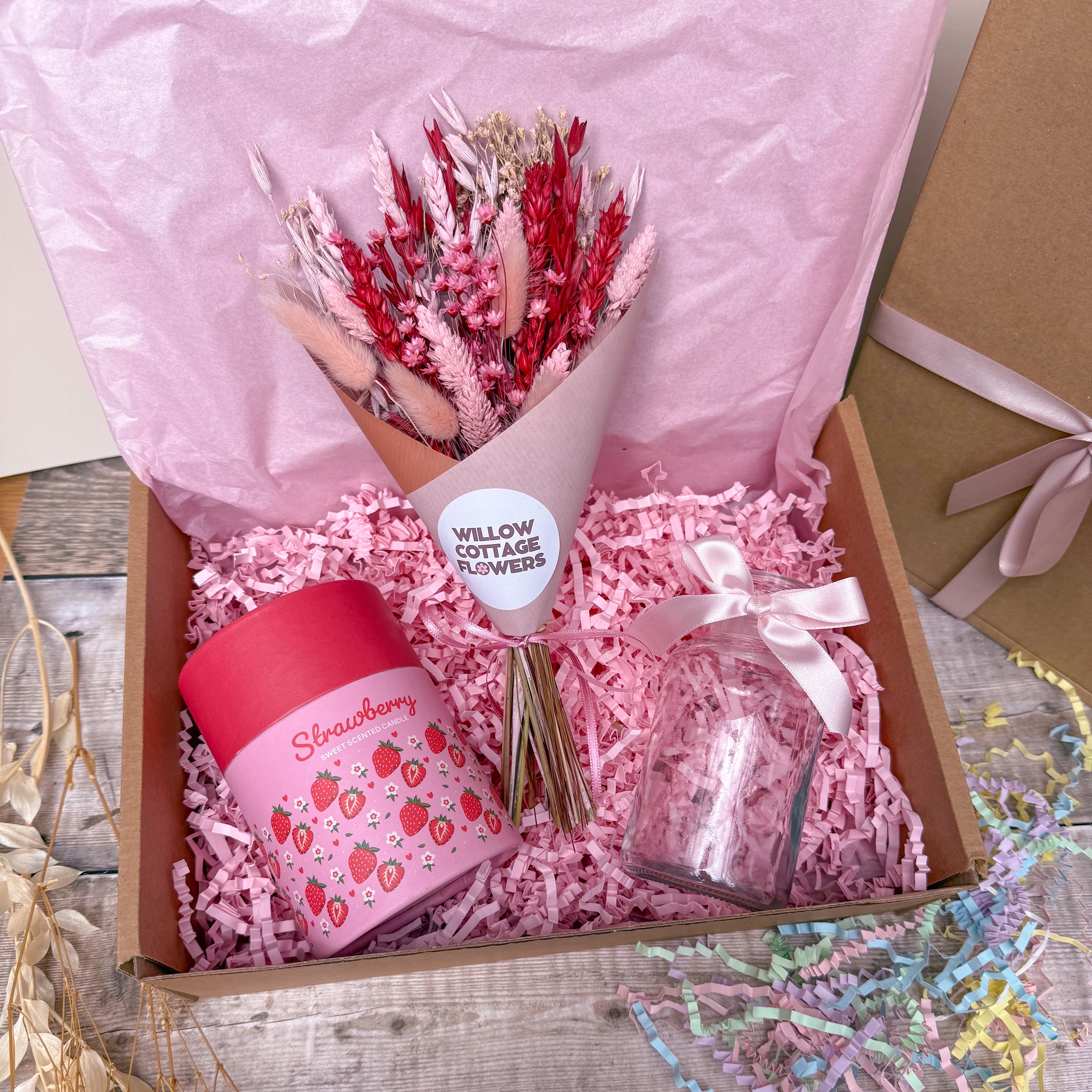 A gift box filled with pink shredded paper contains a bouquet of dried pink and red flowers, a pink strawberry-themed candle, and a small clear glass jar tied with a pale pink ribbon. The box is lined with pink tissue paper and sits on a rustic wooden surface.