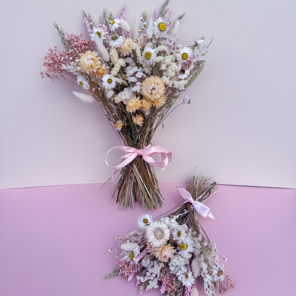 Two dried flower bouquets with white daisies, pink filler flowers, and strawflowers, tied with pink ribbons, displayed against a split light pink and lavender background.
