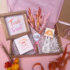 A styled gift box arrangement on a pink surface featuring a framed 'Trick or Treat' sign, a 'Freshly Brewed Pumpkin Spice latte' candle, a white ceramic 'BOO' pumpkin ornament, dried pink and orange flowers and a pumpkin bath bomb in a kraft box with white shredded filler.