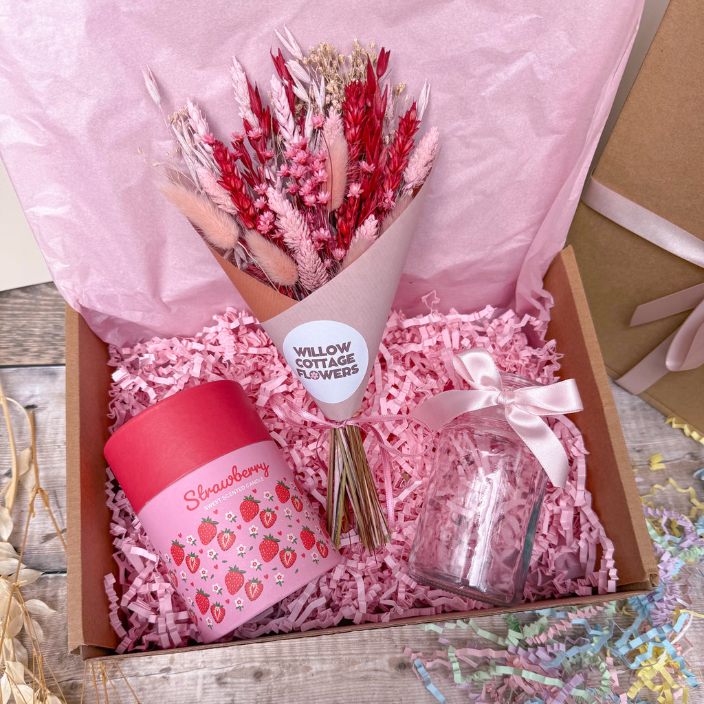 A pink gift box filled with shredded pink paper, containing a bouquet of dried red and pink flowers, a pink strawberry-themed jar candle, and a small clear glass jar tied with a white ribbon. The box is lined with pink tissue paper and sits on a rustic wooden surface.