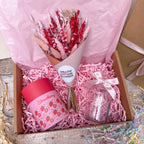 A pink gift box filled with shredded pink paper, containing a bouquet of dried red and pink flowers, a pink strawberry-themed jar candle, and a small clear glass jar tied with a white ribbon. The box is lined with pink tissue paper and sits on a rustic wooden surface.
