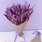 Bouquet of purple and pink flowers wrapped in brown paper with a purple ribbon on a light purple background.