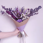 A hand holding a dried flower bouquet wrapped in brown kraft paper and light purple paper, featuring lavender, white daisies, purple eucalyptus, and other dried botanicals against a pale lavender background.