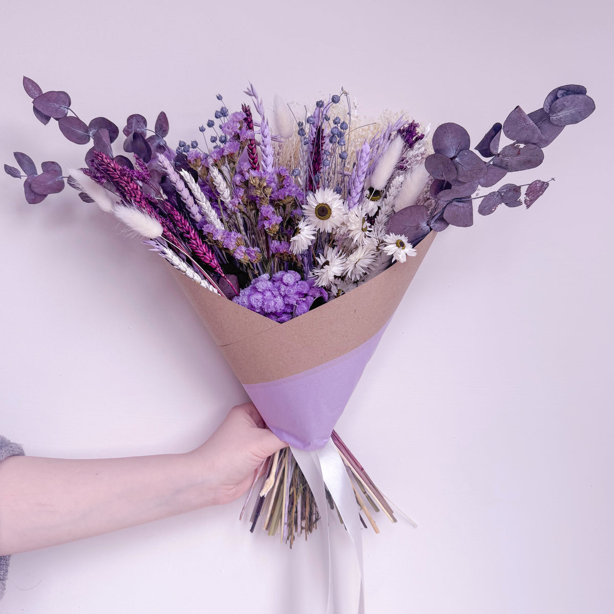 A hand holding a dried flower bouquet wrapped in brown kraft paper and light purple paper, featuring lavender, white daisies, purple eucalyptus, and other dried botanicals against a pale lavender background.