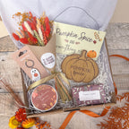 An autumn themed gift box filled with seasonal items including a dried flower bundle, a wooden pumpkin coaster reading 'Pumpkin Spice', a ghost keychain, a round candle tin labeled 'Autumn Spice', a wrapped soap bar, and a cream hanging sign that says 'Pumpkin Spice and everything nice'.