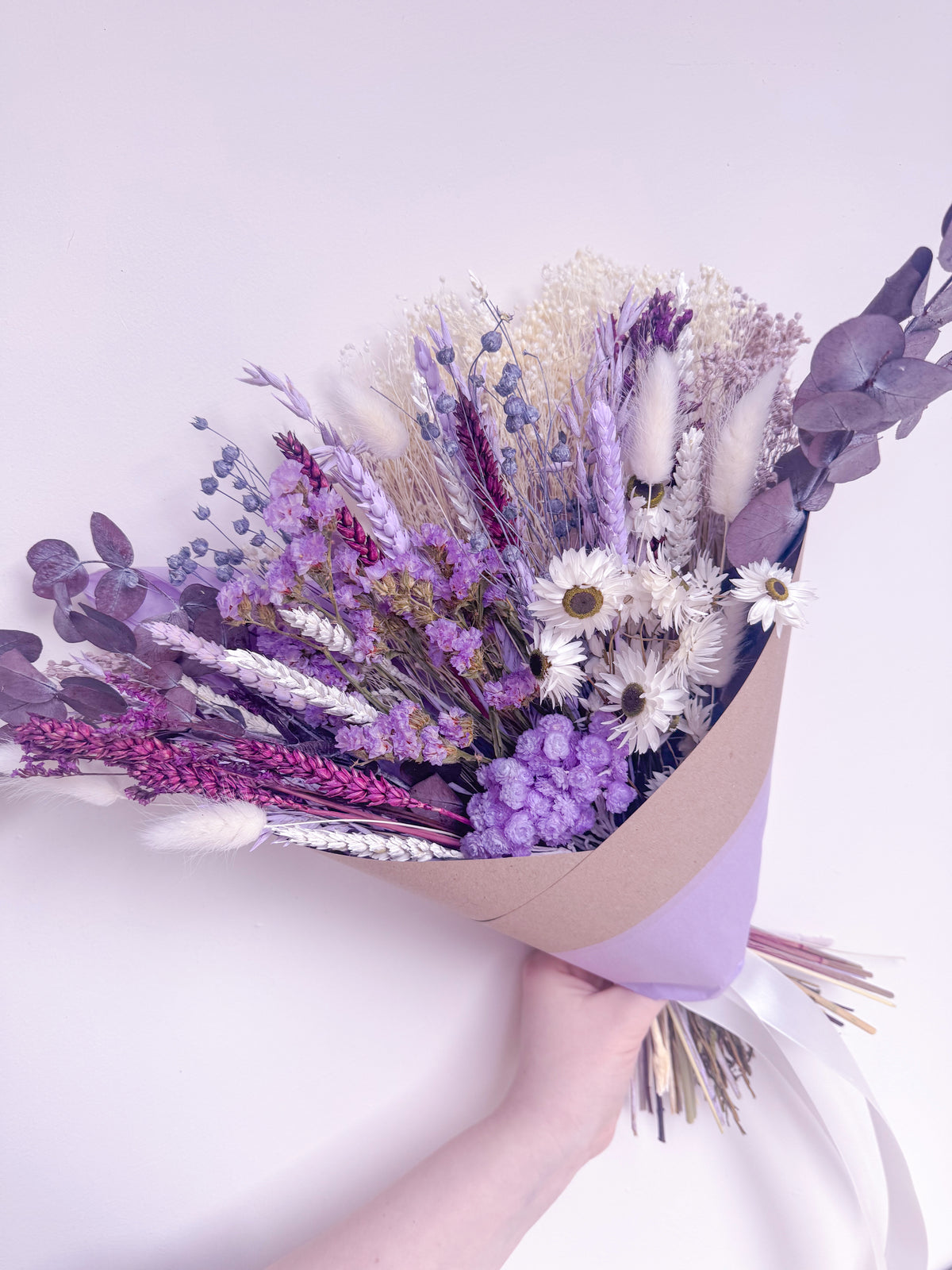 A hand holding a dried flower bouquet wrapped in kraft paper and light purple paper, featuring lavender, purple flowers, white daisies, and soft beige grasses against a pale lavender background.