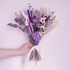 A hand holding a dried flower bouquet with lavender, white daisies, and beige grasses tied with a white ribbon, set against a solid pale pink background.