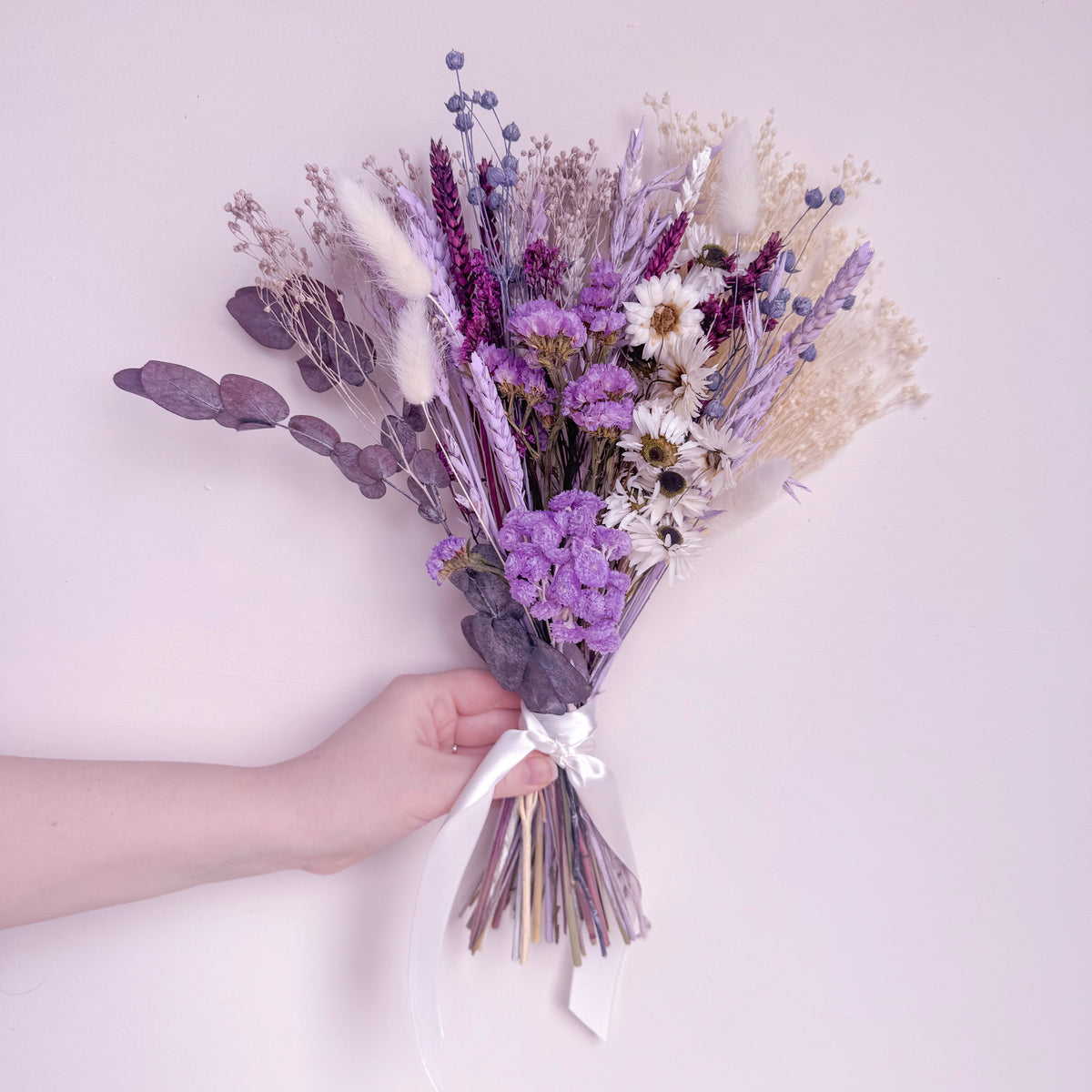 A hand holding a dried flower bouquet with lavender, white daisies, and beige grasses tied with a white ribbon, set against a solid pale pink background.