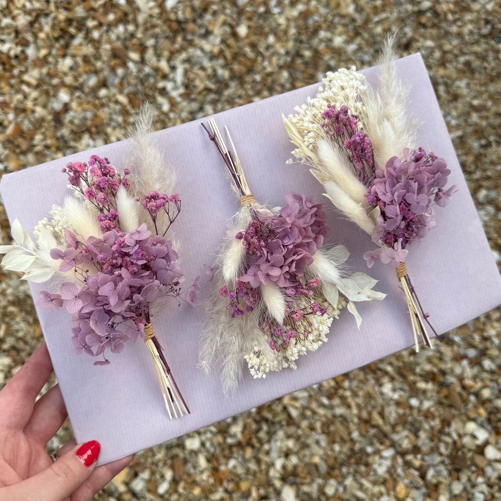 three preserved hydrangea buttonholes with pampas grass and bunny tails, displayed on a pink board