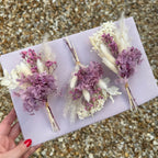 three preserved hydrangea buttonholes with pampas grass and bunny tails, displayed on a pink board