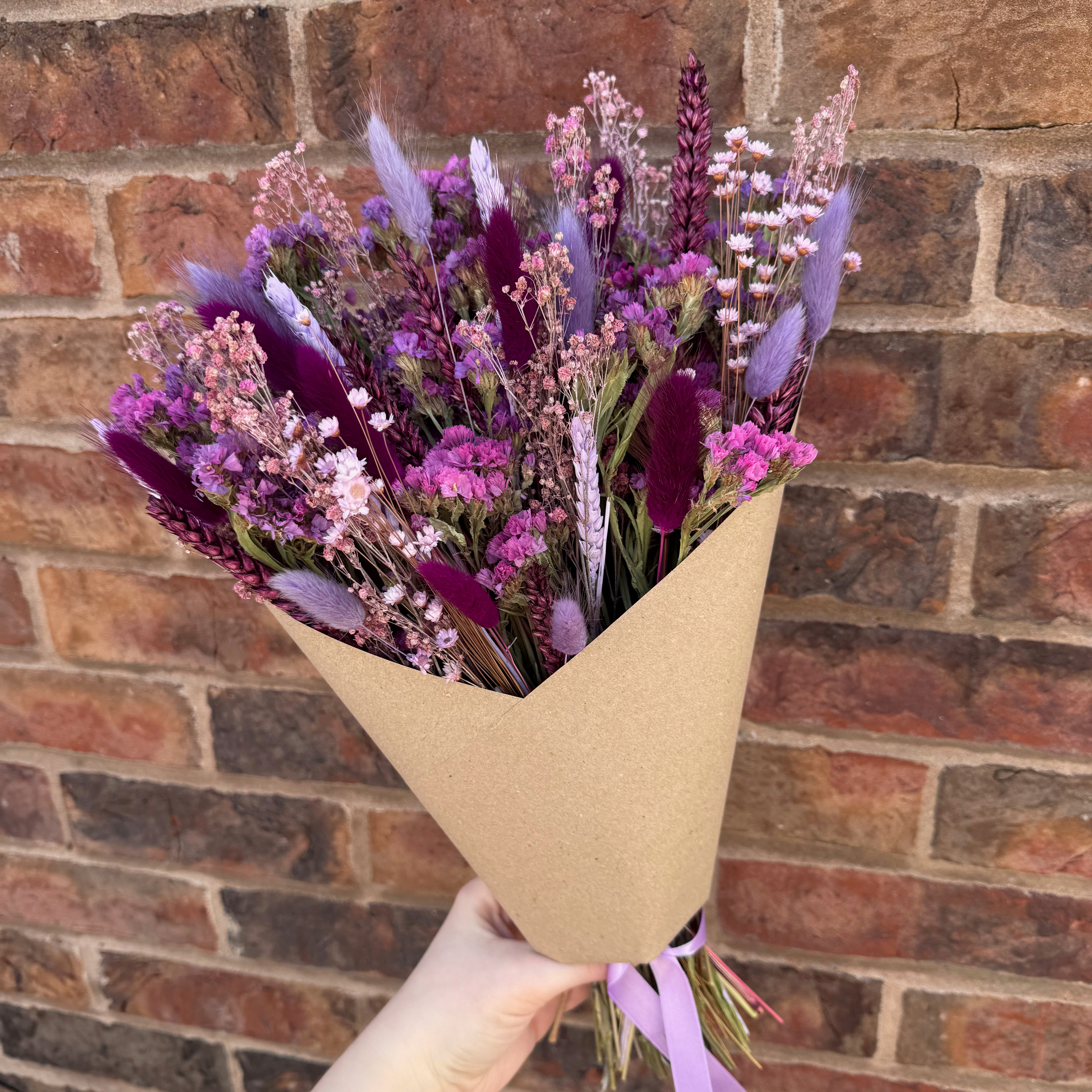 Bouquet of purple and pink flowers wrapped in brown paper held against a brick wall.