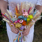 A person in a white dress holds a colorful dried flower bouquet featuring pink, blue, yellow, orange, and beige flowers with pastel ribbons tied at the base.