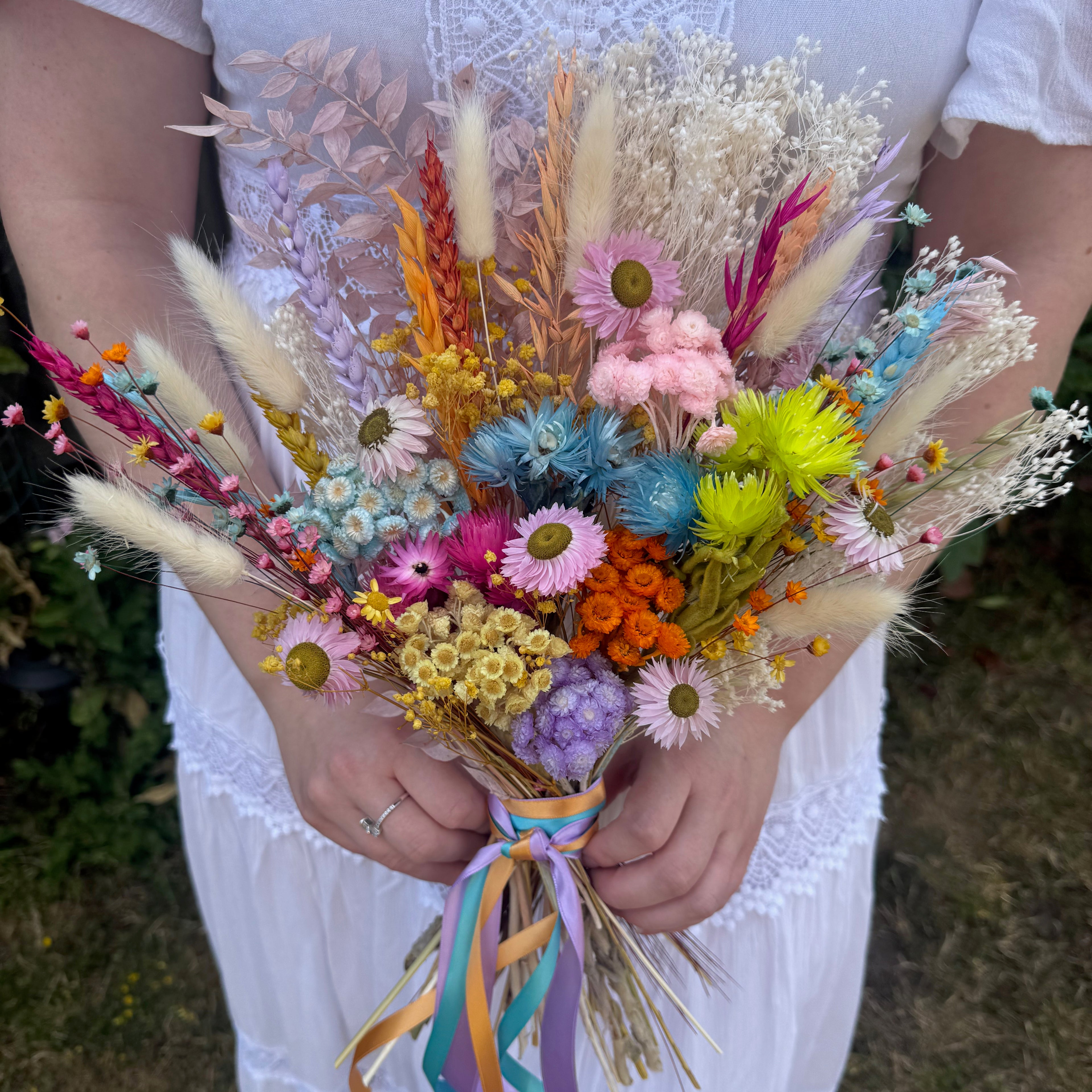A person in a white dress holds a colorful dried flower bouquet featuring pink, blue, yellow, orange, and beige flowers with pastel ribbons tied at the base.