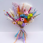 A vibrant, multicolored dried flower bouquet featuring bunny tails, daisies, and various wildflowers tied with pastel blue, purple, and orange ribbons, set against a plain light grey background.
