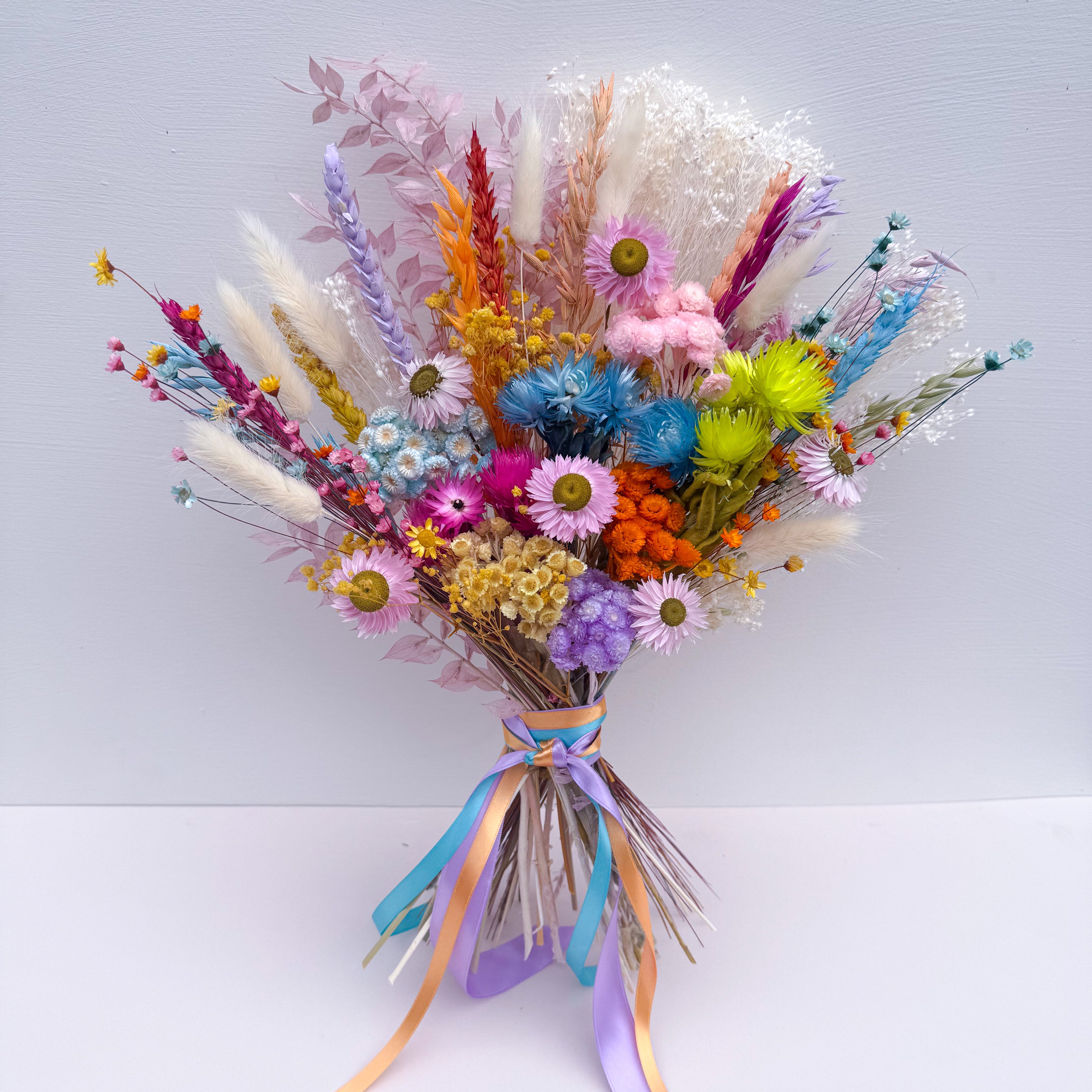 A vibrant, multicolored dried flower bouquet featuring bunny tails, daisies, and various wildflowers tied with pastel blue, purple, and orange ribbons, set against a plain light grey background.