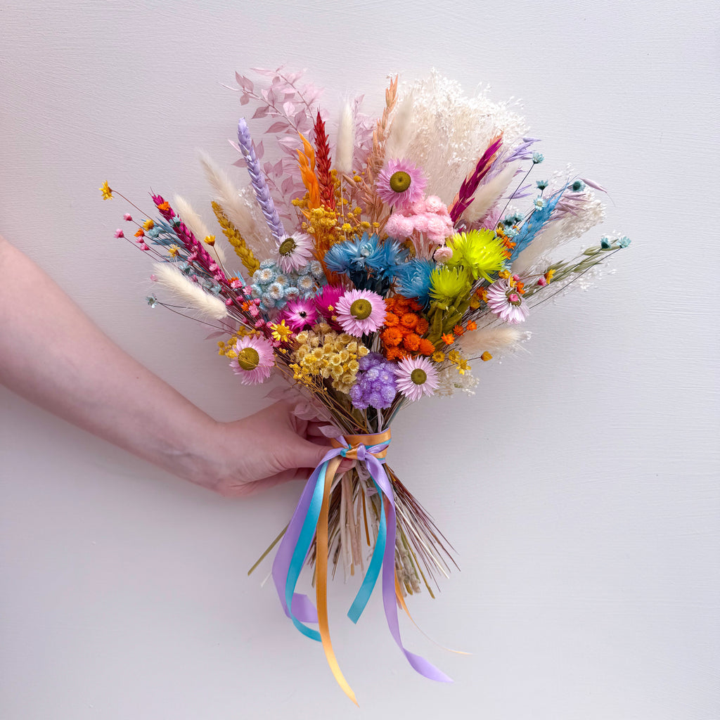 A hand holding a colorful dried flower bouquet with pastel and bright blooms, daisies and long blue and yellow ribbons against a plain light background.