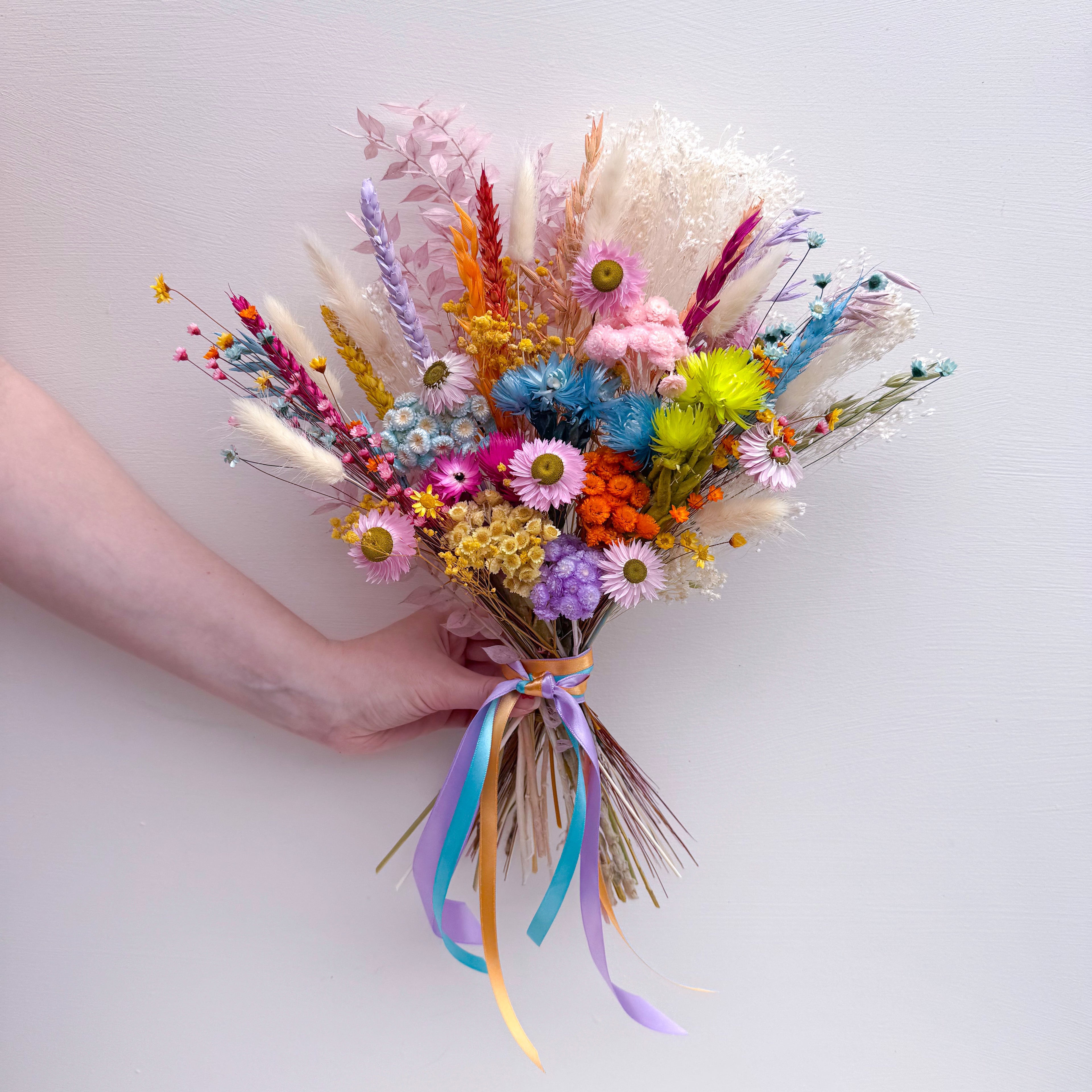 A hand holding a colorful dried flower bouquet with pastel and bright blooms, daisies and long blue and yellow ribbons against a plain light background.