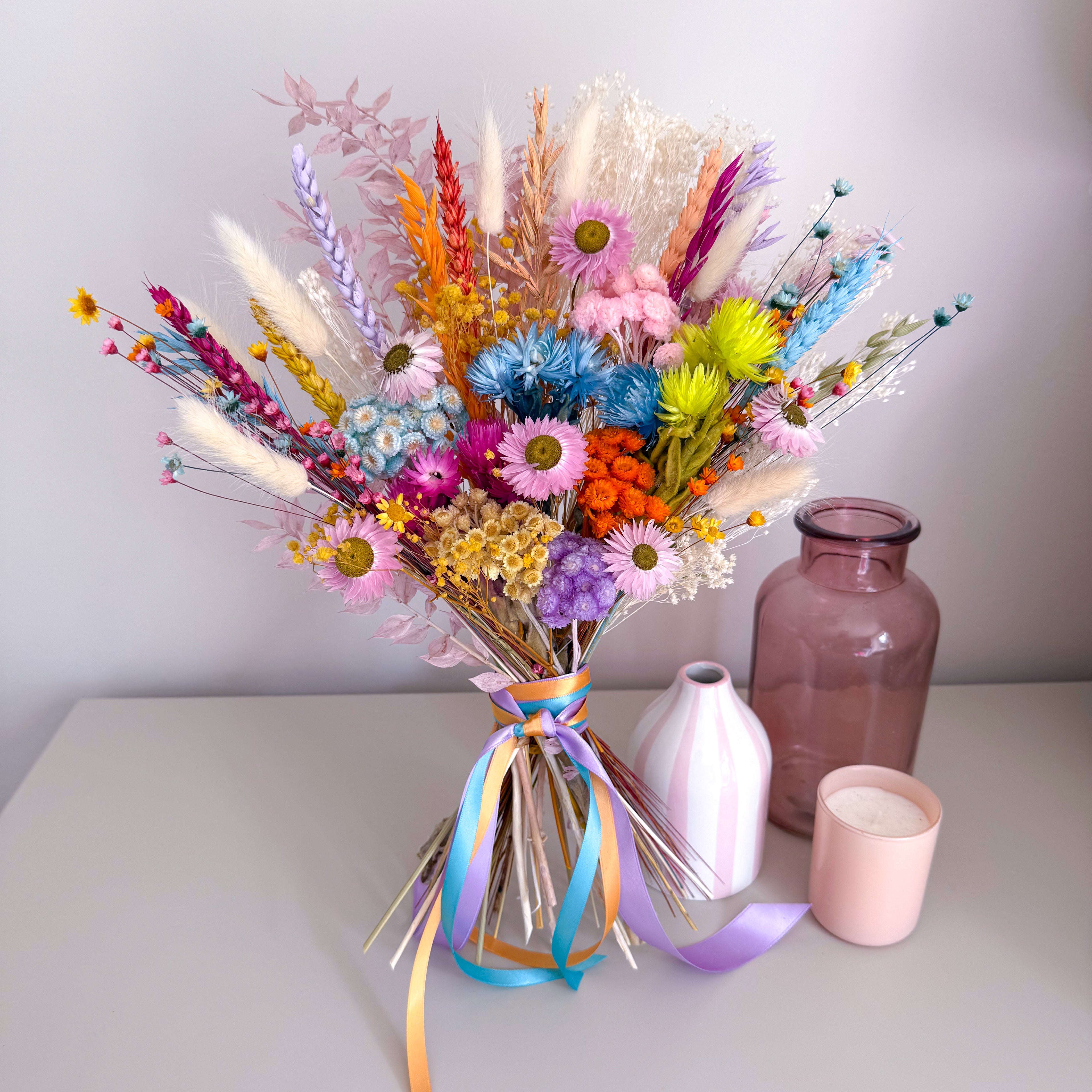 A colorful dried flower bouquet in a clear glass vase with pastel ribbons, styled on a tabletop beside a pink glass bottle and a pink candle against a light gray wall.