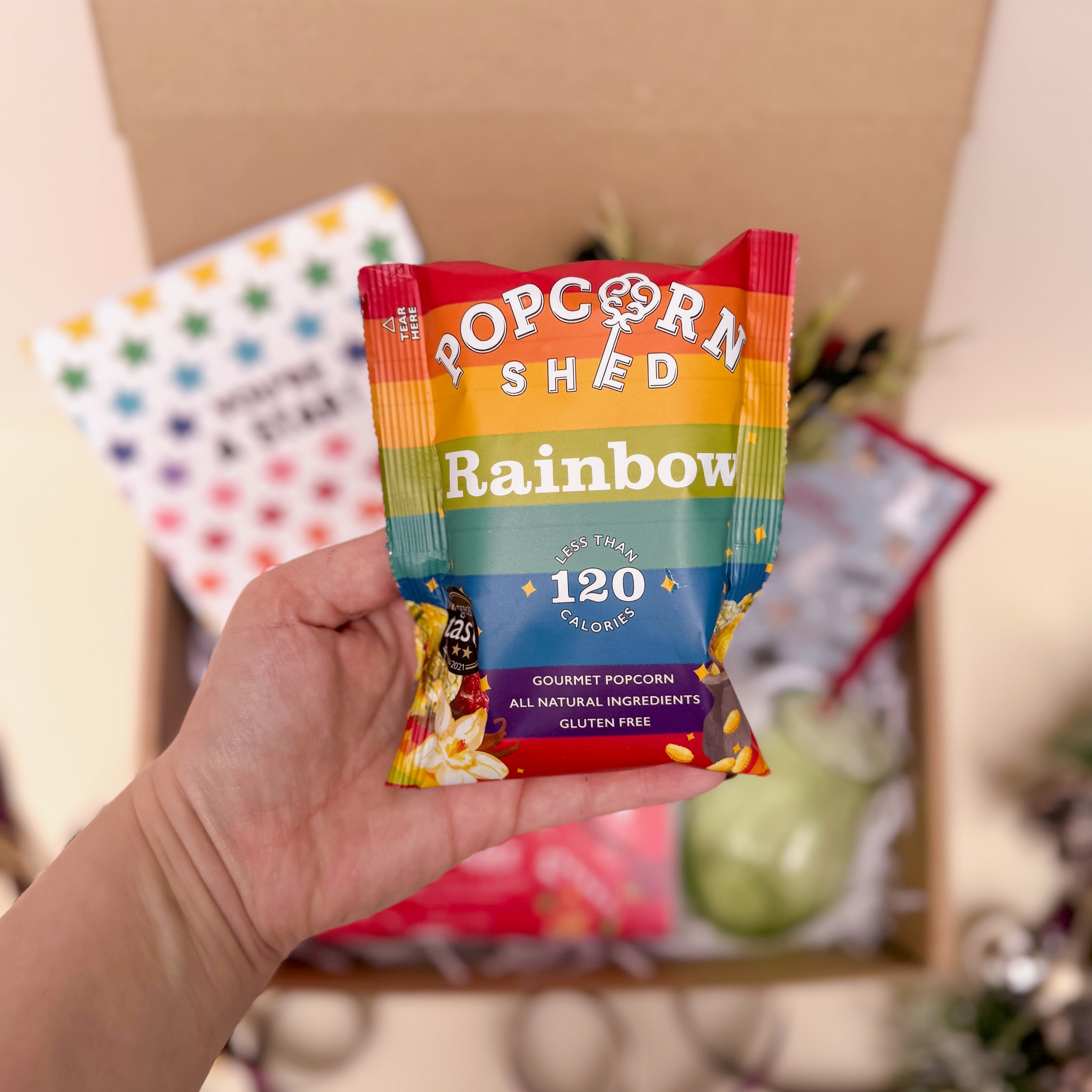 Hand holding a Popcorn Shed Rainbow popcorn packet with a blurred background