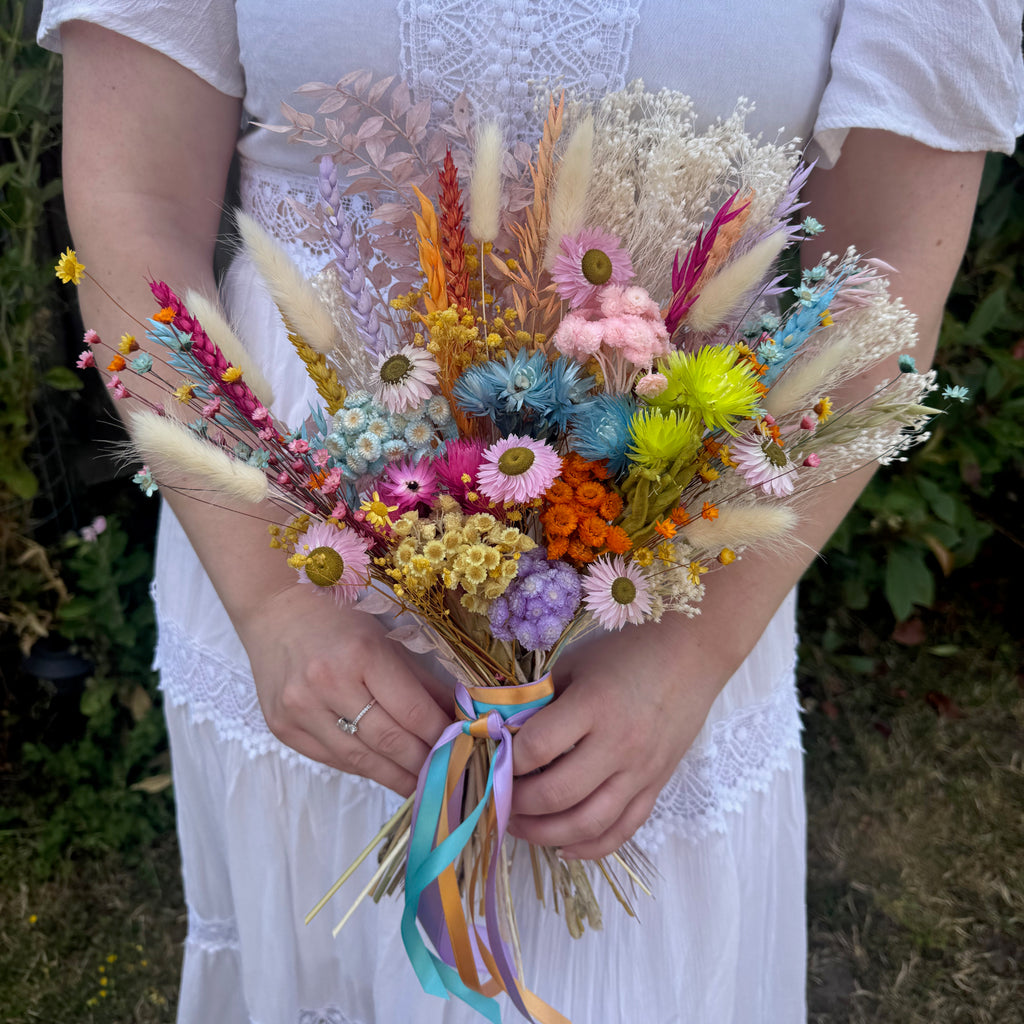 A person in a white wedding dress holds a colourful rainbow dried flower bouquet outdoors. The bouquet includes pink, yellow, orange, blue, purple, and beige wildflowers and grasses tied with pastel ribbons.