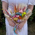 A person in a white wedding dress holds a colourful rainbow dried flower bouquet outdoors. The bouquet includes pink, yellow, orange, blue, purple, and beige wildflowers and grasses tied with pastel ribbons.