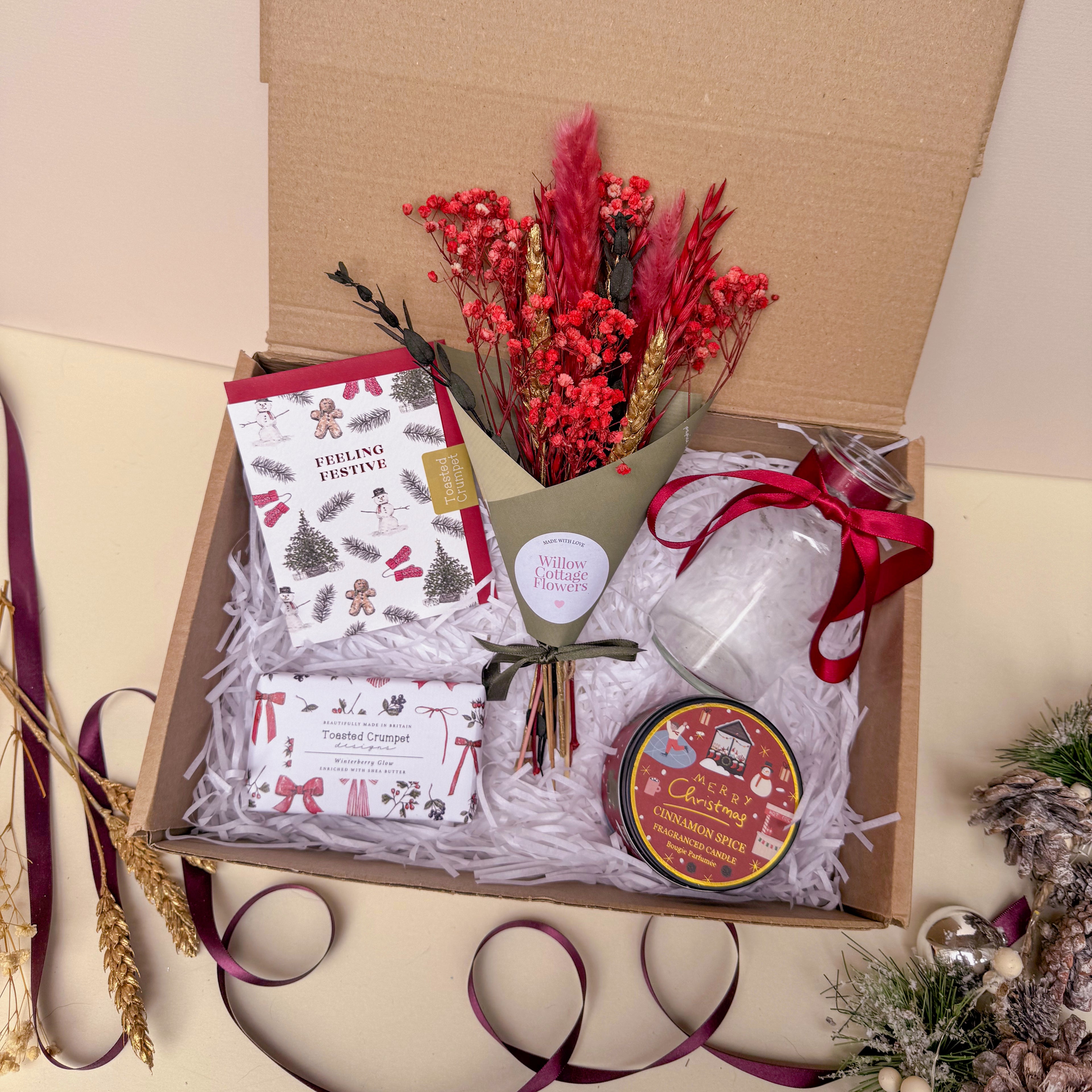 Gift box with festive items including a bouquet, cards, and a candle on a beige background.
