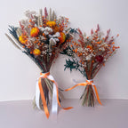 Two dried flower bouquets with orange, yellow, white, and green botanical elements, tied with white and orange ribbons, set against a plain light gray background.