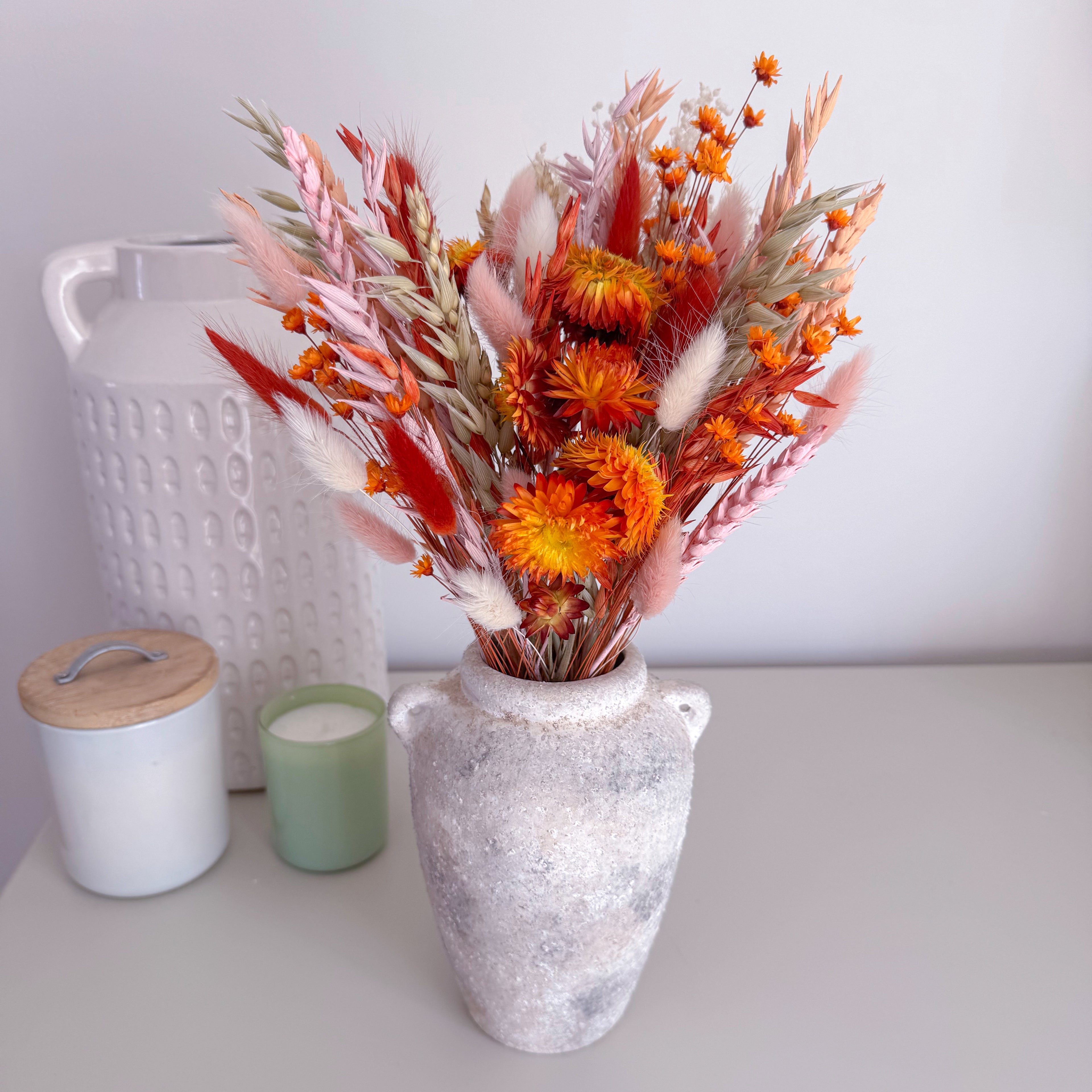 A dried floral arrangement in a textured grey ceramic vase, styled on a tabletop with a white textured canister, a green candle, and a white jar with a wooden lid in the background.