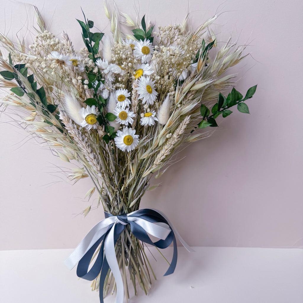 A dried floral arrangement featuring white daisies with yellow centers, oat grass, wheat, and green ruscus, tied with a green and white ribbon, set against a soft pink and white background.