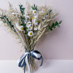 A dried floral arrangement featuring white daisies with yellow centers, oat grass, wheat, and green ruscus, tied with a green and white ribbon, set against a soft pink and white background.