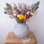 A rustic grey ceramic vase filled with a colourful dried flower arrangement, displayed on a wooden block against a plain light background.