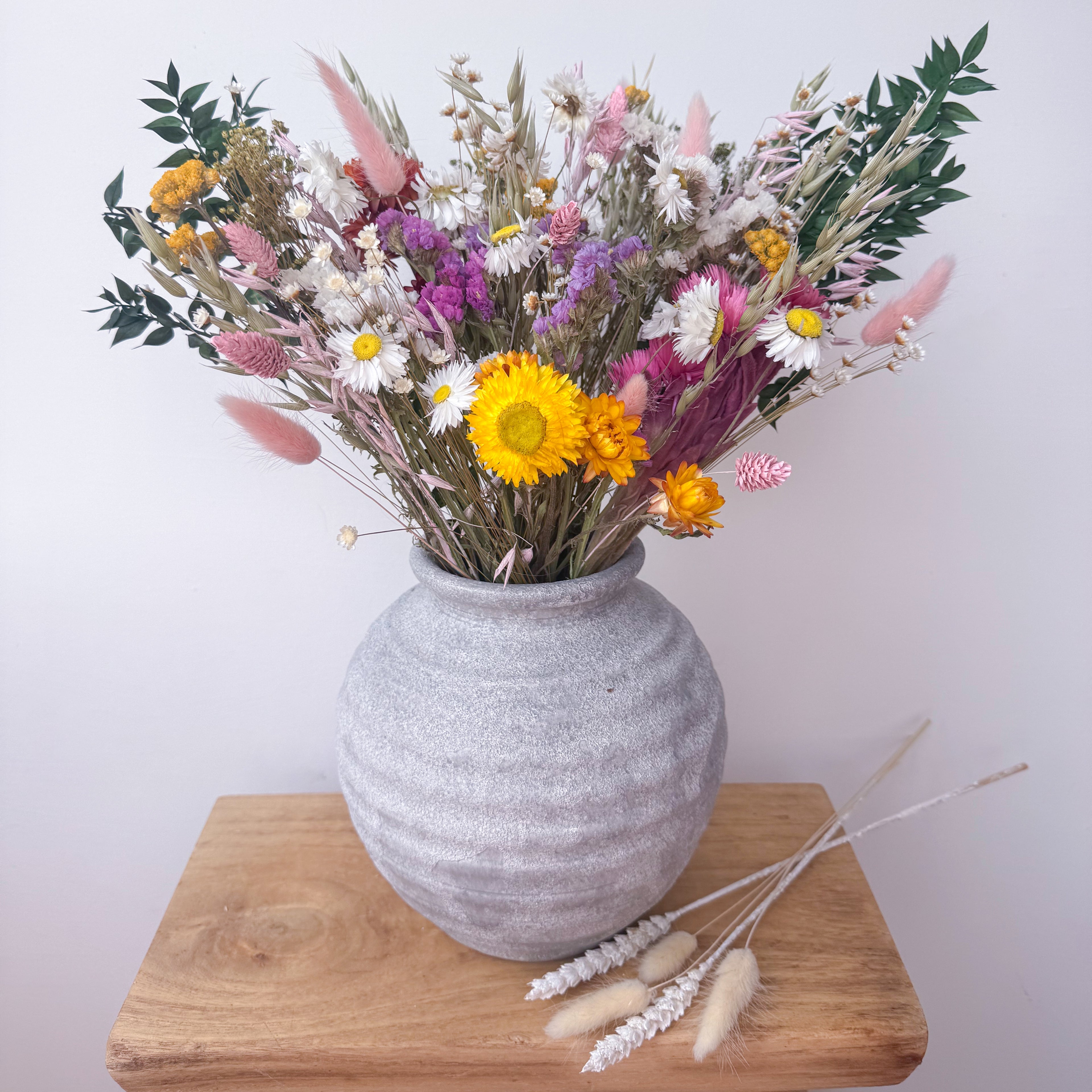 A rustic grey ceramic vase filled with a colourful dried flower arrangement, displayed on a wooden block against a plain light background.
