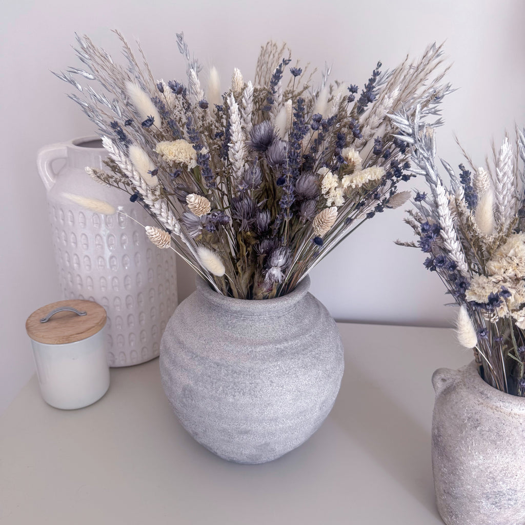 A round textured grey ceramic vase filled with dried lavender, bunny tails and wheat stems, styled on a tabletop next to a white textured ceramic jug and a small white jar with a wooden lid.