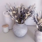 A round textured grey ceramic vase filled with dried lavender, bunny tails and wheat stems, styled on a tabletop next to a white textured ceramic jug and a small white jar with a wooden lid.
