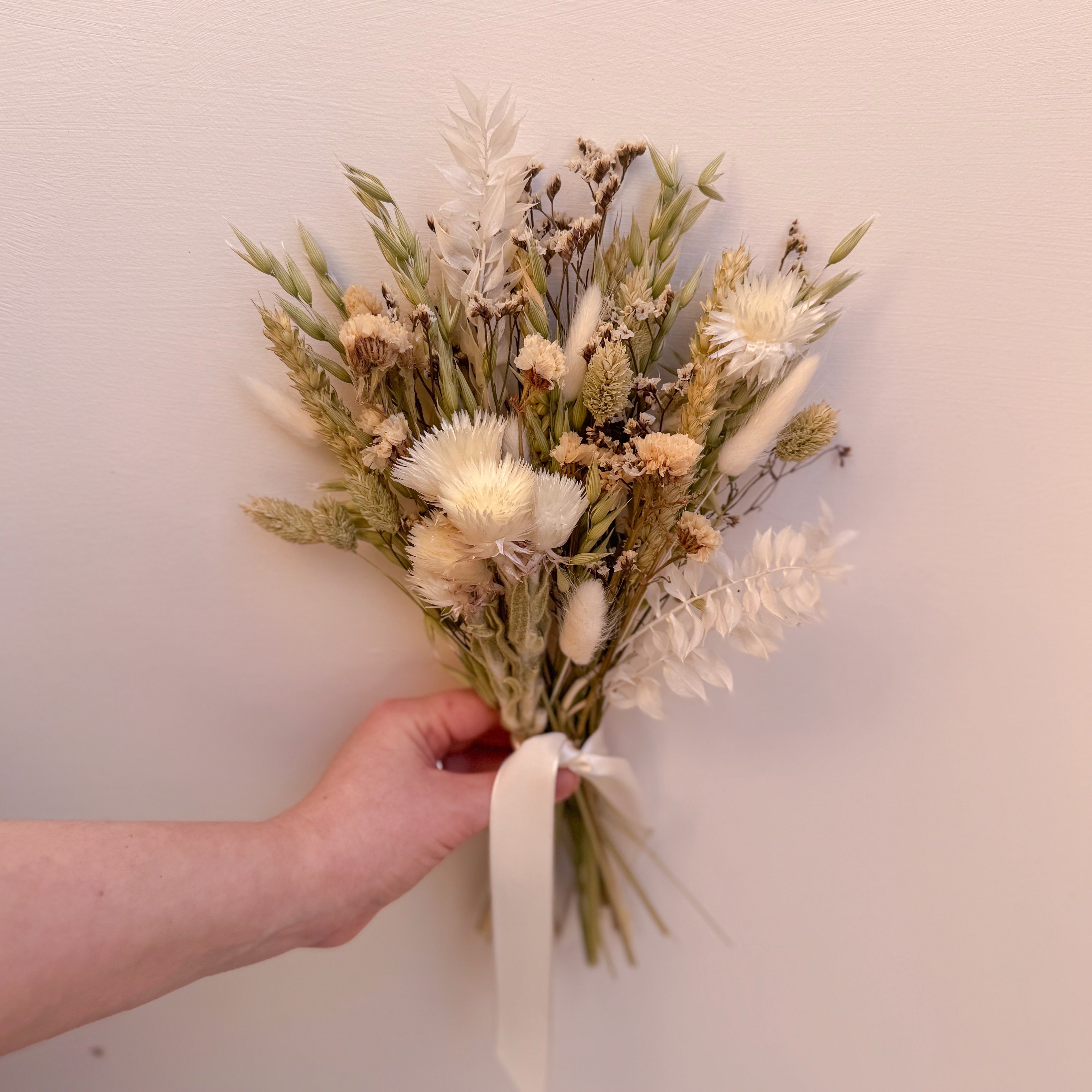 Hand holding a small bouquet of green and white dried flowers with a plain background