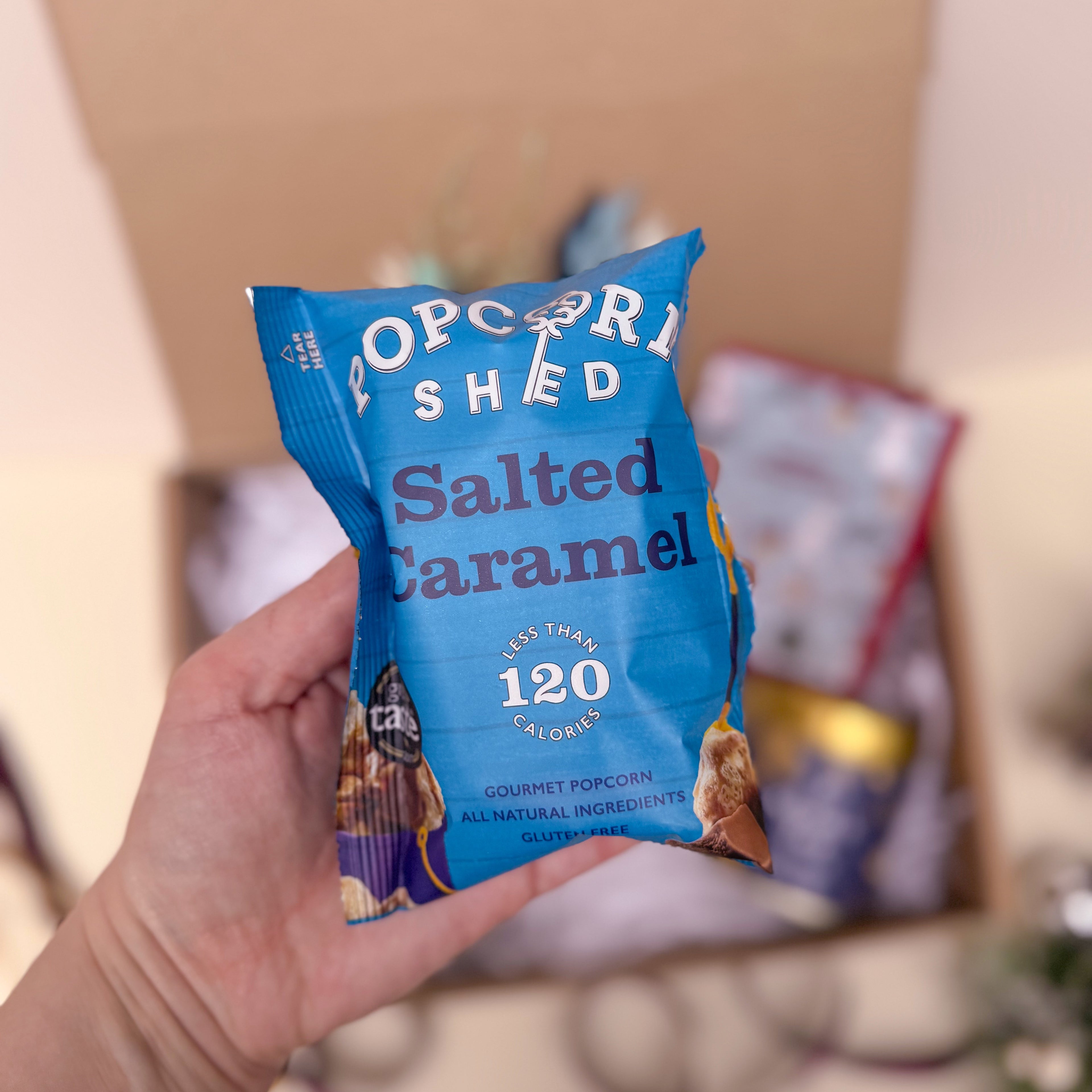 Hand holding a blue 'Popcorn Shed Salted Caramel' popcorn packet with a blurred background.